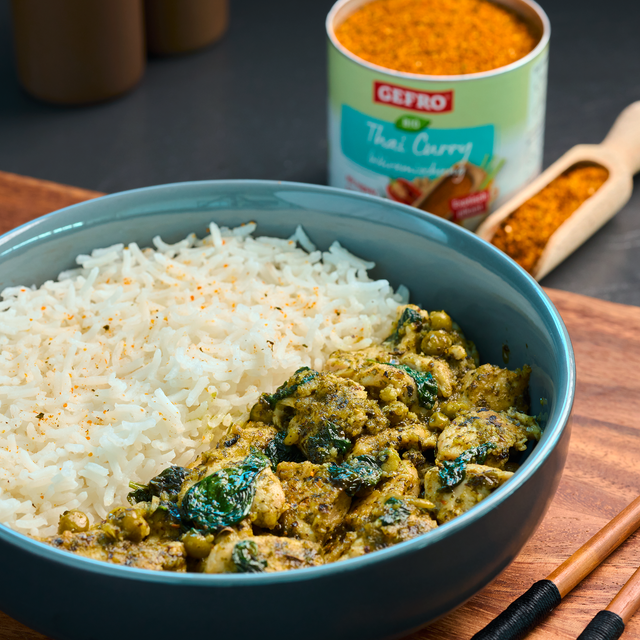 Schale mit weißem Reis und grünem Thai-Curry mit Gemüse, daneben Dose mit Gewürz und Holzlöffel auf Tisch