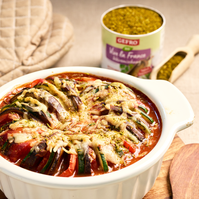 Überbackenes Gemüse in Tomatensauce in einer weißen Auflaufform, daneben Kräutergewürz und Holzlöffel.