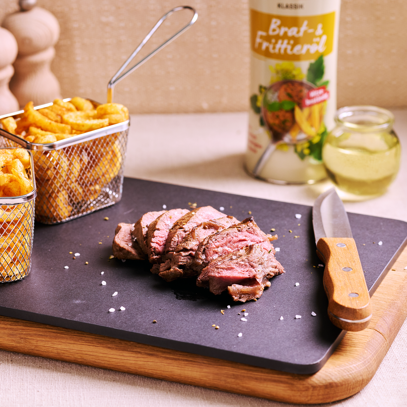 Aufgeschnittenes Steak auf schwarzer Platte mit Messer, Pommes in Körben und Brat- und Frittieröl im Hintergrund