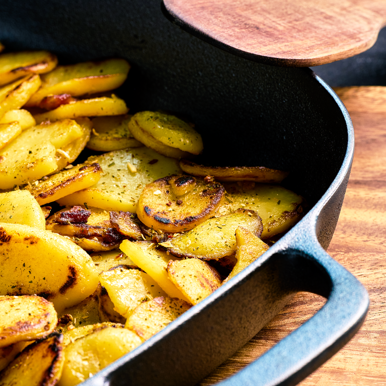 Gebratene Kartoffelscheiben in schwarzer Pfanne mit Holzdeckel, servierfertig auf Holztisch angerichtet