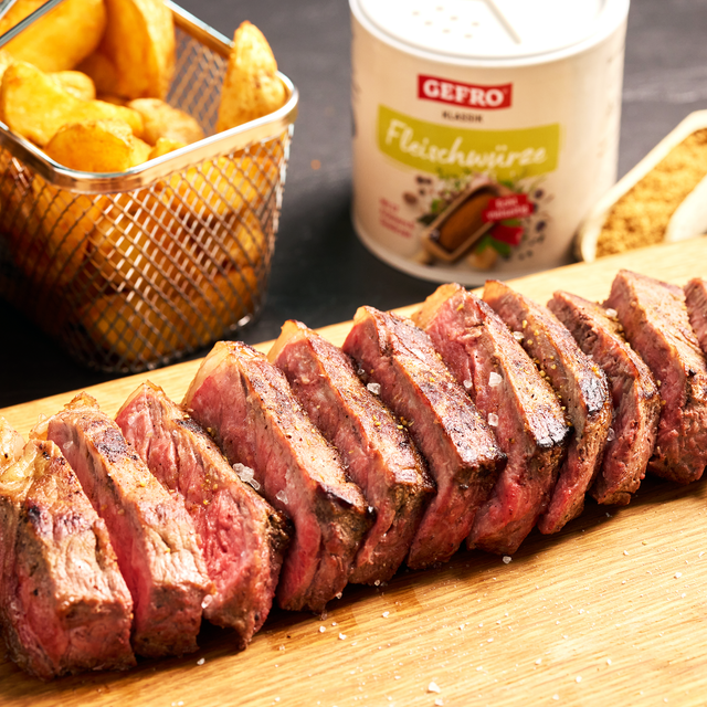 Aufgeschnittenes Steak auf Holzbrett mit grobem Salz, dazu Kartoffelecken und Gefro Fleischwürze im Hintergrund.