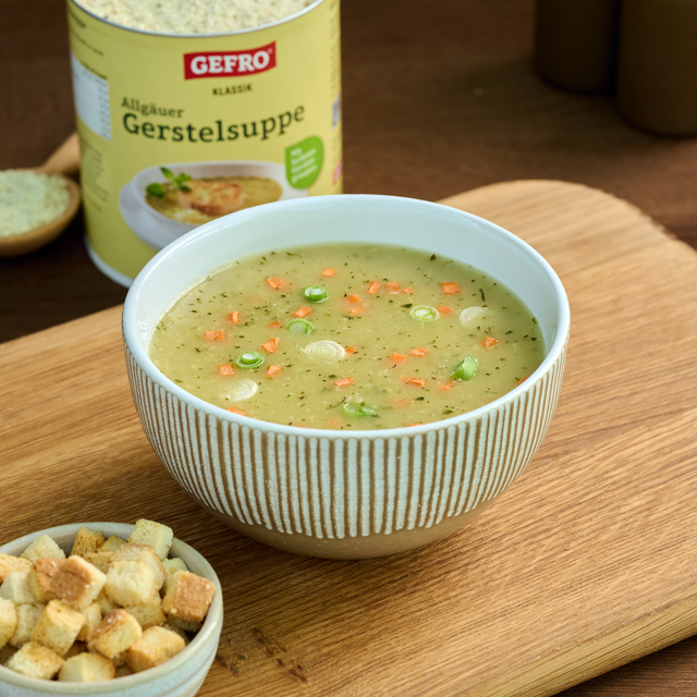 Schüssel mit Allgäuer Gerstelsuppe auf Holzbrett, dazu Croutons; die Gefro Verpackung steht im Hintergrund.