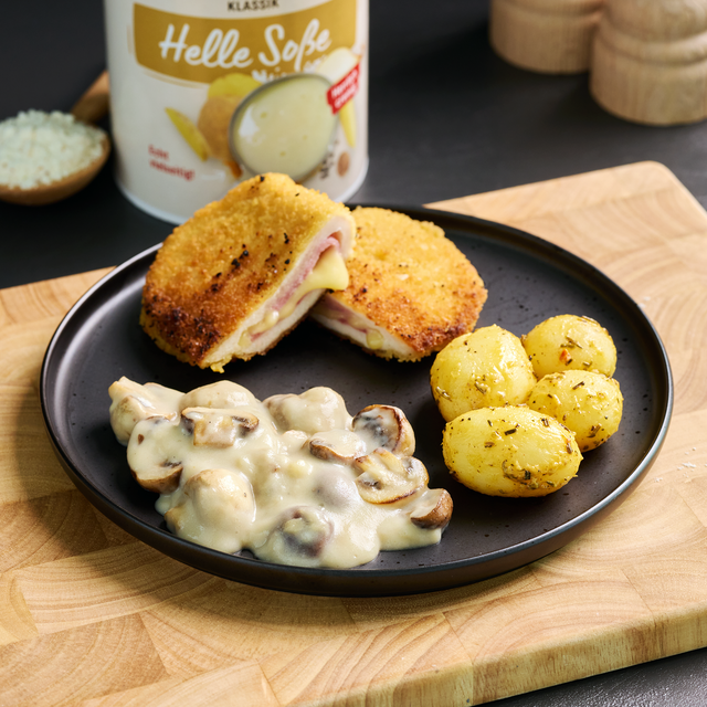 Cordon bleu mit Kräutersoße, Champignons und kleinen Kartoffeln auf schwarzem Teller, servierfertig angerichtet