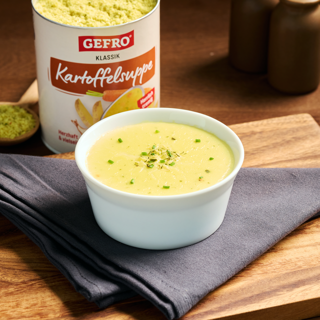 Schale mit cremiger Kartoffelsuppe auf grauem Tuch, dahinter Dose GEFRO Klassik Kartoffelsuppe auf Holztisch