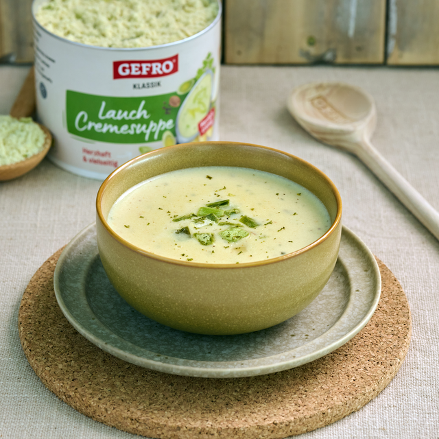 Schüssel mit heller Lauchcremesuppe auf Teller, daneben GEFRO Dose und Holzlöffel auf hellem Tisch.