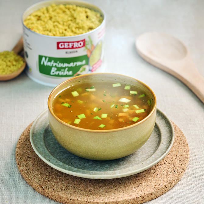 Schale mit klarer Gemüsebrühe auf Teller, dahinter offene Dose Gefro Naturwürze Brühe und ein Holzlöffel.