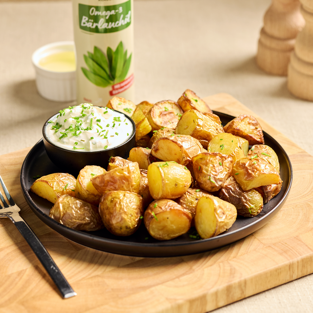 Gebratene Kartoffelhälften auf schwarzem Teller mit Kräuterdip, passend zu Omega-3-Bärlauchöl im Hintergrund