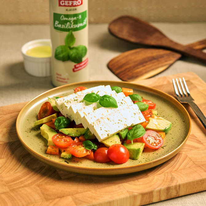 Salat mit Feta, Avocado, Tomaten und Basilikum auf Teller, daneben Basilikumwürze und Besteck auf Holzbrett