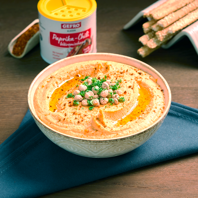 Schüssel mit cremigem Hummus, Kichererbsen und Öl, dahinter Paprika-Chili-Würzmischung und Grissini