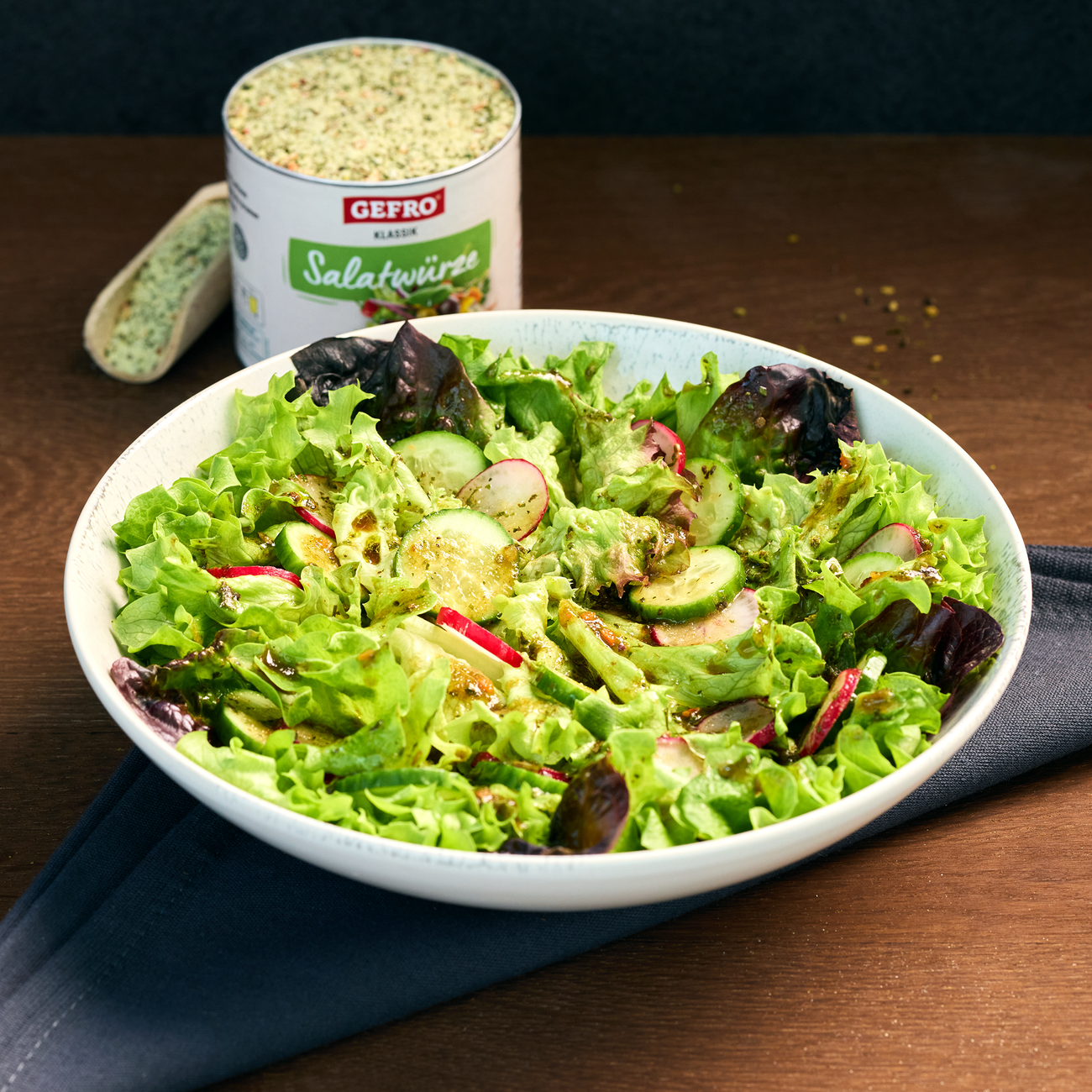 Schüssel mit frischem Blattsalat, Gurken und Radieschen, im Hintergrund eine Dose GEFRO Salatwürze auf dem Tisch