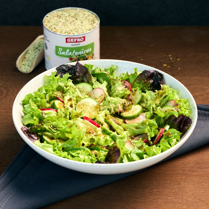 Schüssel mit frischem Blattsalat, Gurken und Radieschen, im Hintergrund eine Dose GEFRO Salatwürze auf dem Tisch