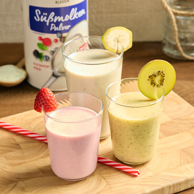 Drei Smoothies in Gläsern mit Erdbeere, Kiwi und Banane, daneben eine Dose Süßmolkenpulver auf Holzbrett.