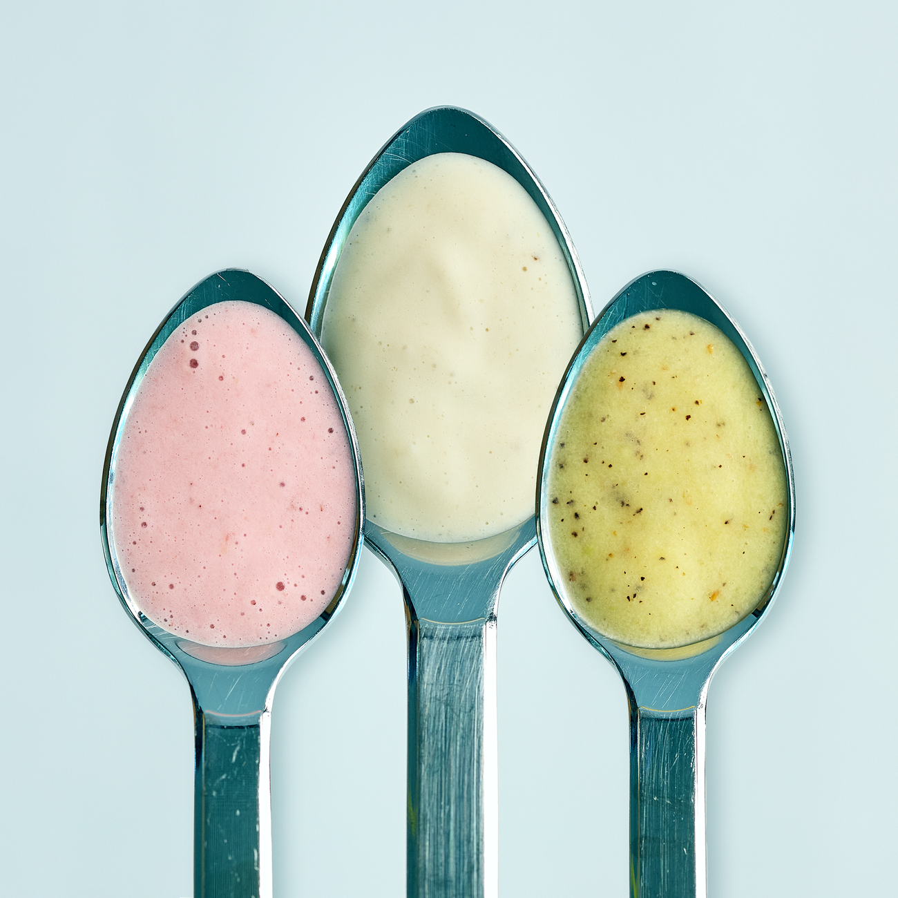 Drei Metalllöffel mit rosa, weißer und gelbgrüner Creme vor hellem Hintergrund, übersichtliche Darstellung verschiedener Sorten