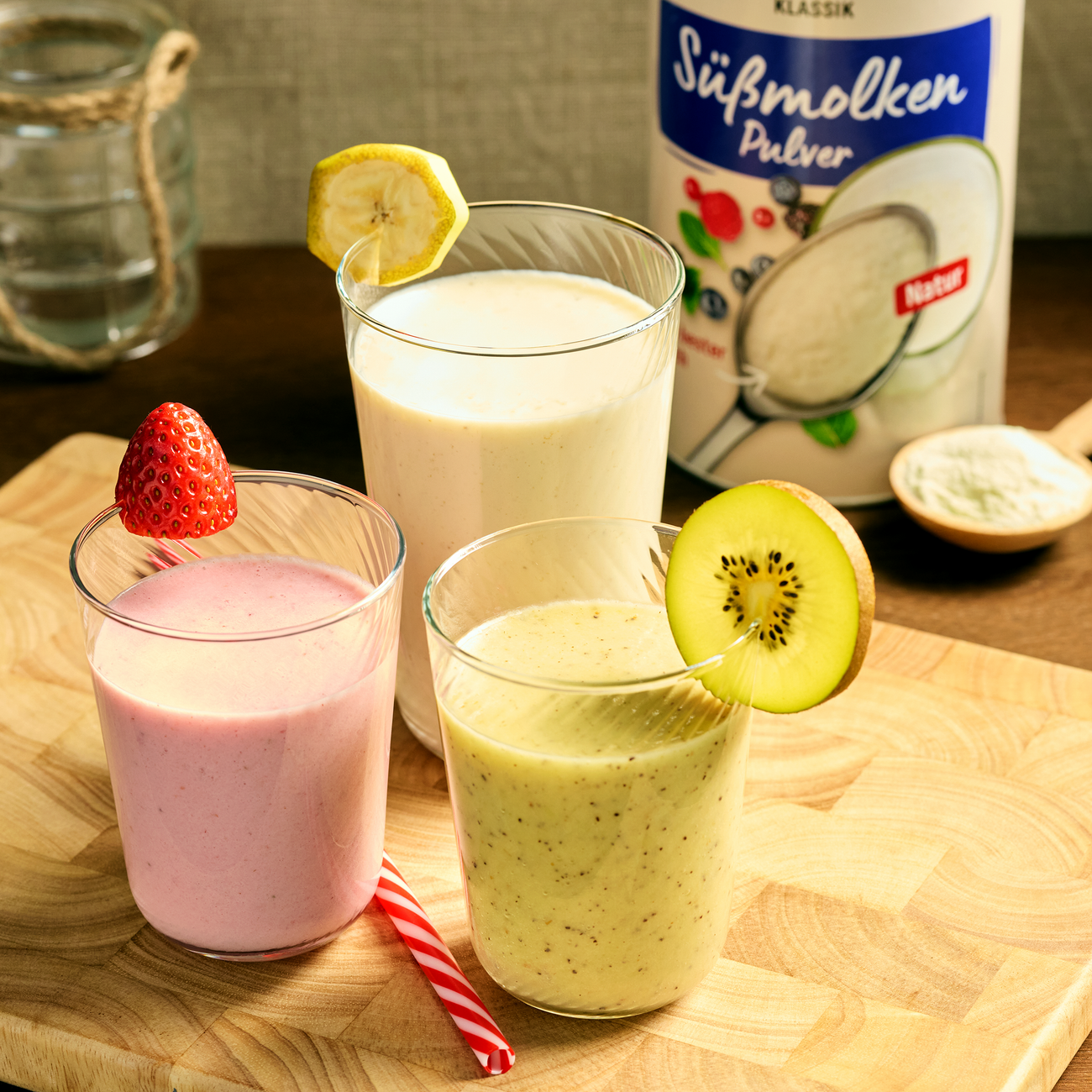 Drei Smoothies in Gläsern mit Erdbeere, Kiwi und Banane, daneben Süßmolkenpulver auf einem Holzbrett.