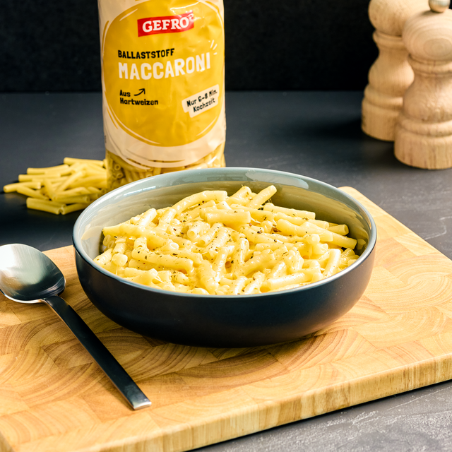 Schale mit cremigen Maccheroni auf Holzbrett, daneben Löffel und Packung GEFRO Ballaststoff Maccaroni im Hintergrund