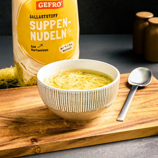 Schale mit klarer Nudelsuppe auf Holzbrett, daneben Löffel und Packung Gefro Ballaststoff-Suppennudeln