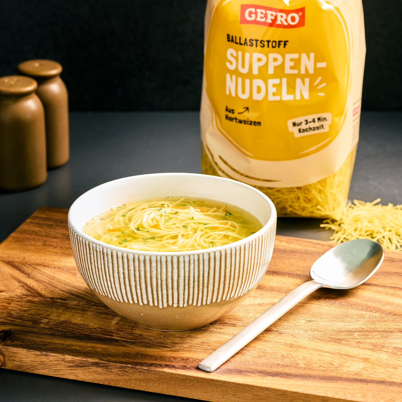 Schale mit klarer Nudelsuppe auf Holzbrett, daneben Löffel und Packung GEFRO Suppennudeln im Hintergrund