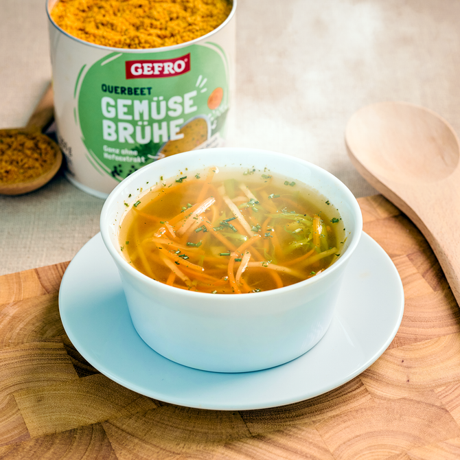 Schale mit klarer Gemüsesuppe auf Untertasse, daneben Dose GEFRO Querbeet Gemüsebrühe und Holzlöffel.