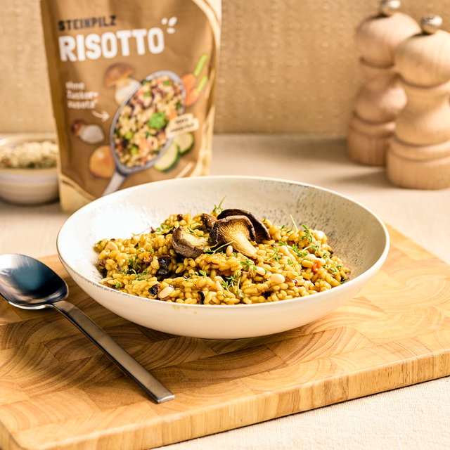 Schale mit Pilz-Risotto auf Holzbrett, garniert mit Kräutern und Pilzen, Packung im Hintergrund sichtbar