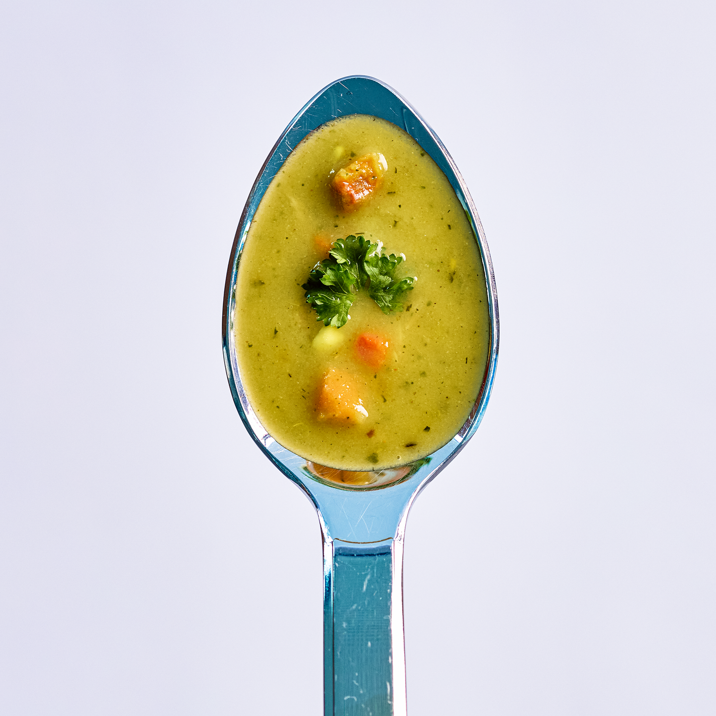 Löffel mit cremiger Gemüsesuppe, kleinen Gemüsewürfeln und Petersilie als Garnitur vor hellem Hintergrund