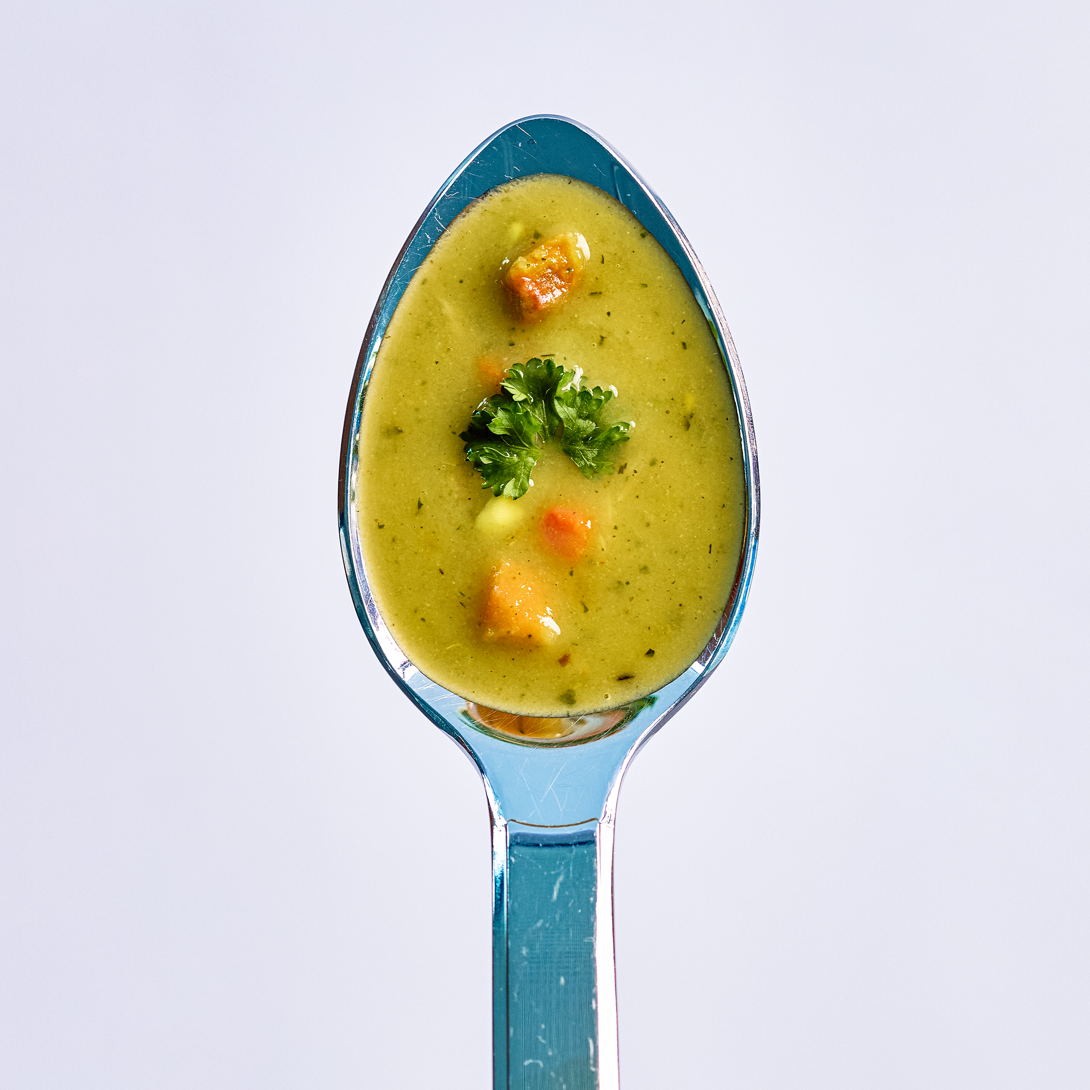 Löffel mit cremiger Gemüsesuppe, kleinen Gemüsewürfeln und Petersilie als Garnitur vor hellem Hintergrund