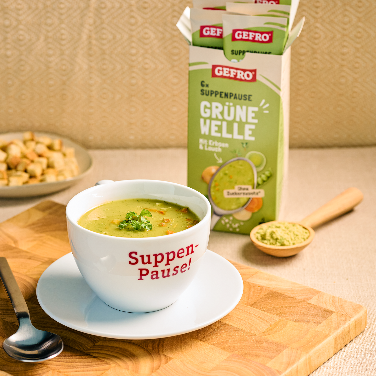 Tasse mit grüner Erbsen-Lauch-Suppe vor geöffneter GEFRO-Packung und Löffel mit Suppenpulver auf dem Tisch