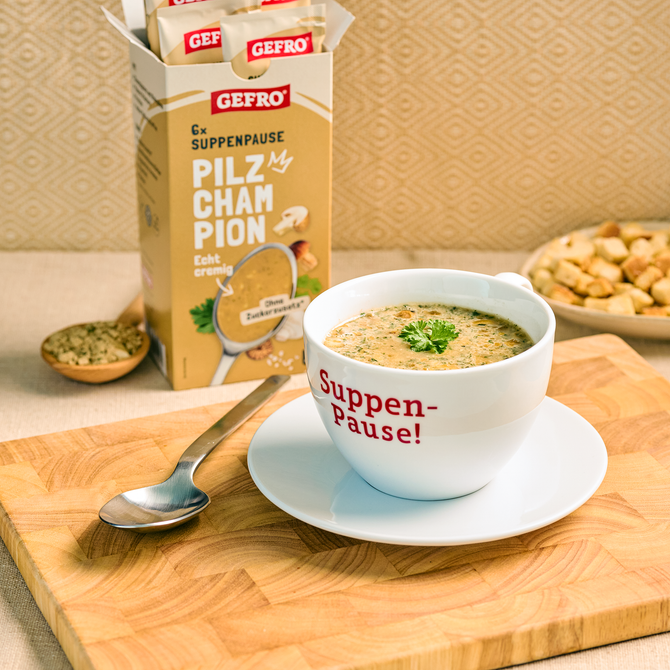 Tasse mit cremiger Pilzsuppe auf Unterteller, daneben GEFRO Suppenpause Pilz Champignon und ein Löffel.