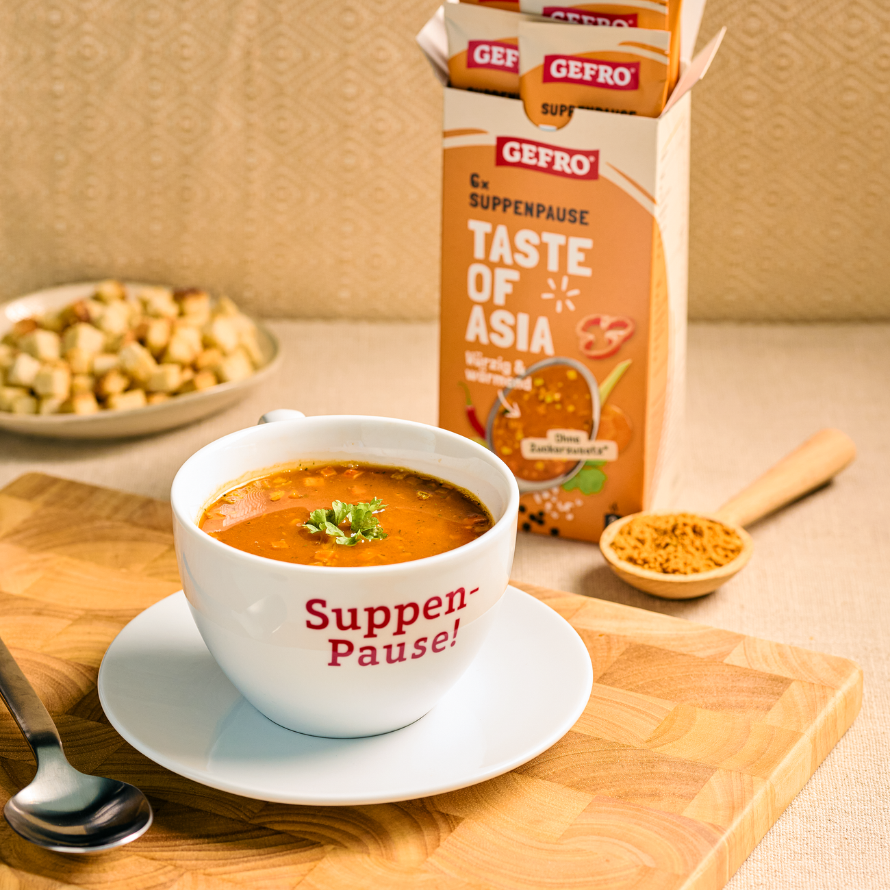 Schale mit orangefarbener Asia-Suppe vor GEFRO Suppenpause Taste of Asia und Gewürzlöffel auf Holzbrett