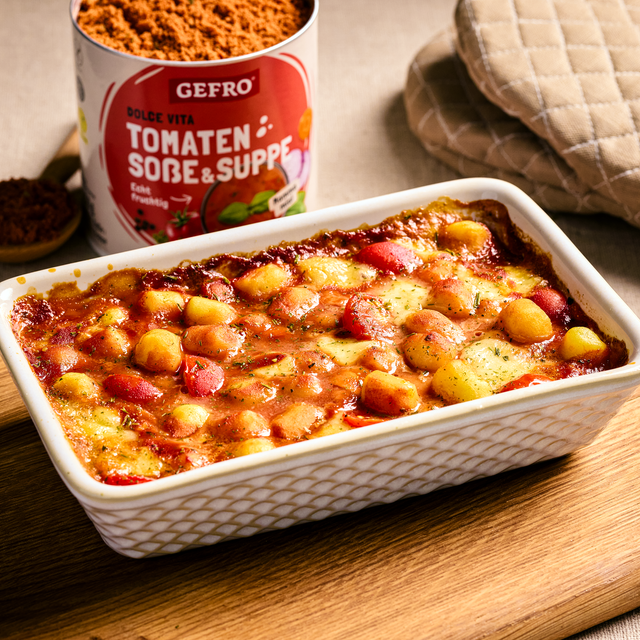 Auflauf mit Gnocchi, Tomatensoße und geschmolzenem Käse in einer weißen Form, daneben Gewürzdose auf Holzbrett