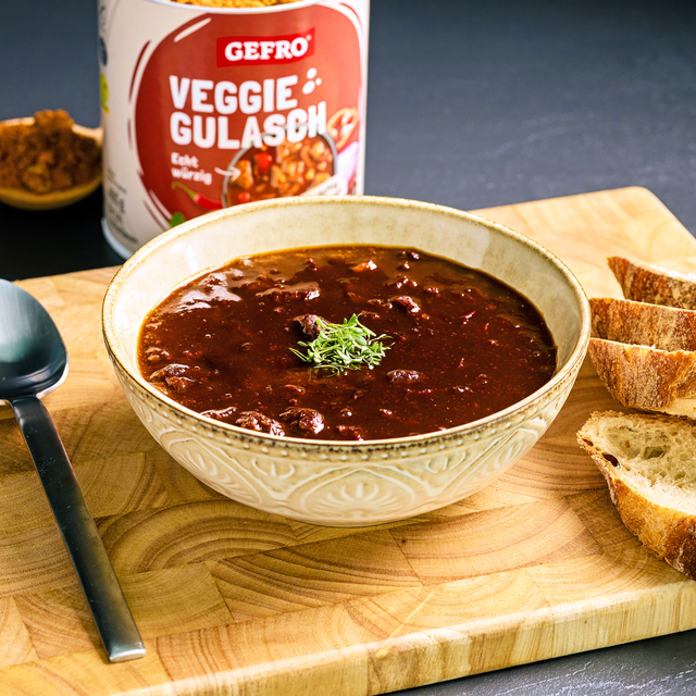 Schüssel mit dunklem Veggie-Gulasch auf Holzbrett, dazu Brotscheiben; im Hintergrund die Packung von Gefro.