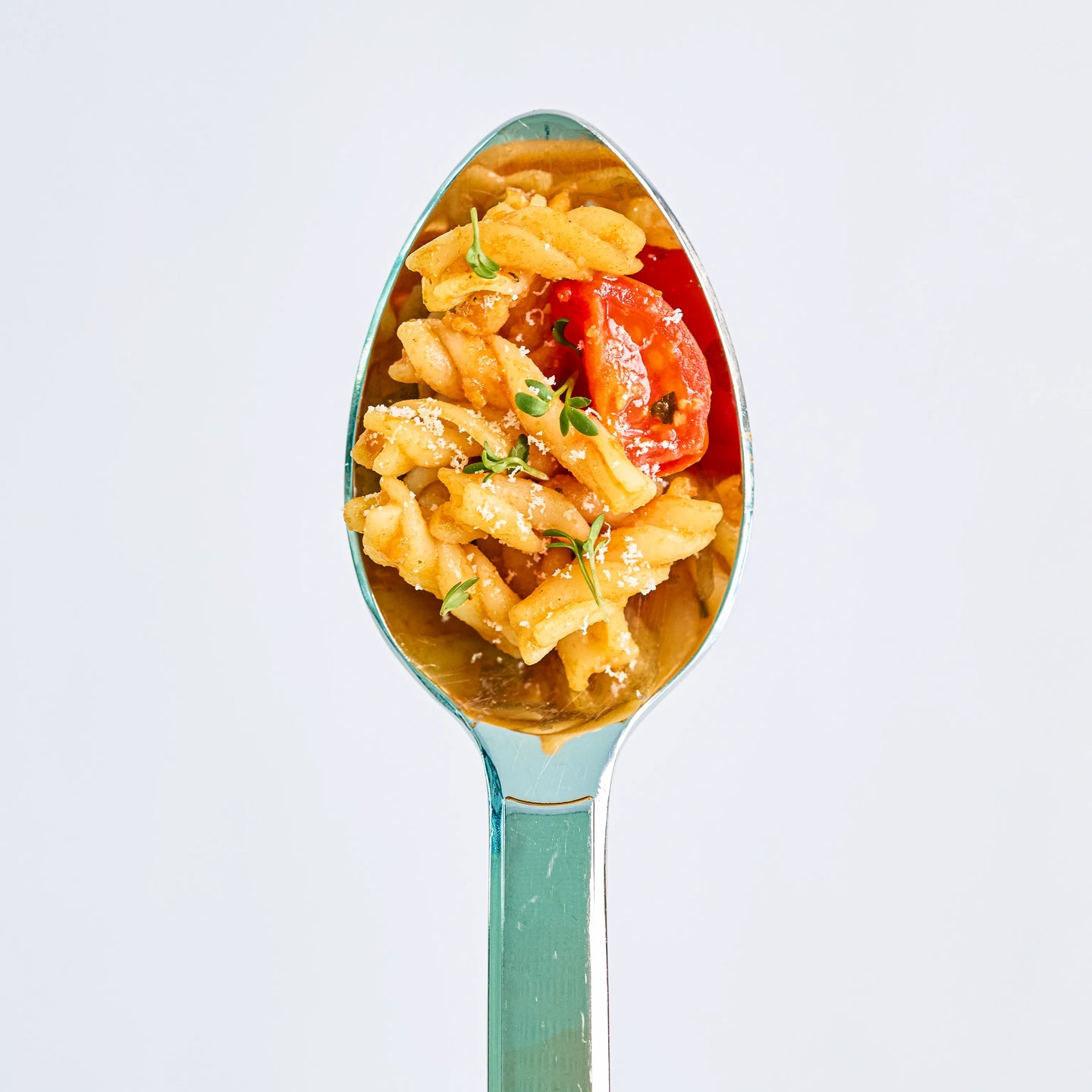 Löffel mit Fusilli-Nudeln, Tomatenscheibe, Kräutern und Käse, appetitlich angerichtet vor hellem Hintergrund