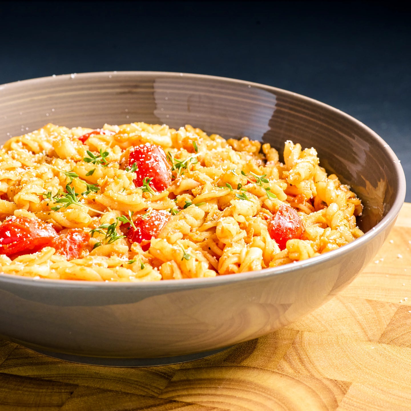 Graue Schüssel mit Fusilli in Tomatensauce, Kirschtomaten, geriebenem Käse und frischen Kräutern auf Holzbrett