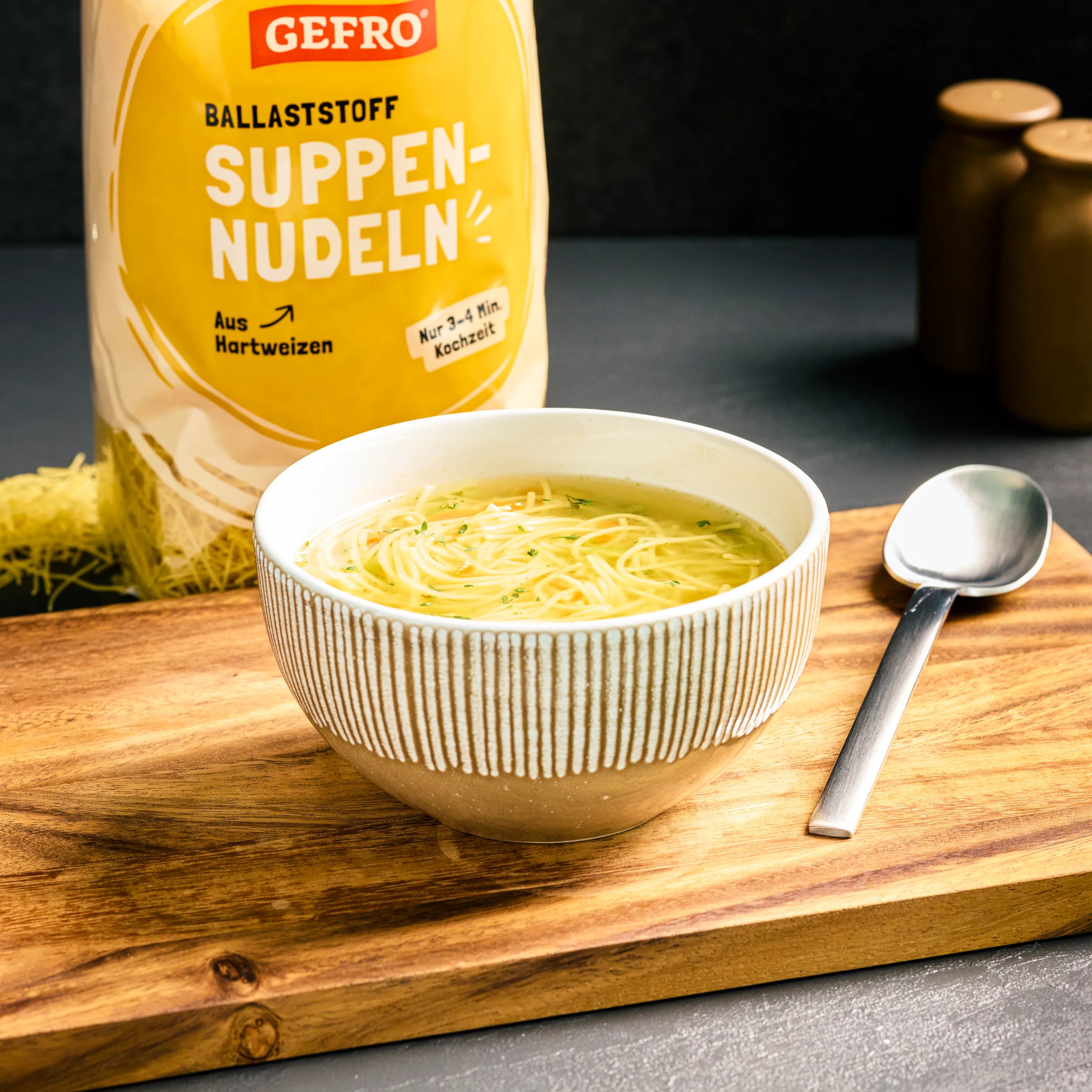 Schale mit klarer Nudelsuppe auf Holzbrett, daneben Löffel und Packung GEFRO Ballaststoff Suppennudeln.