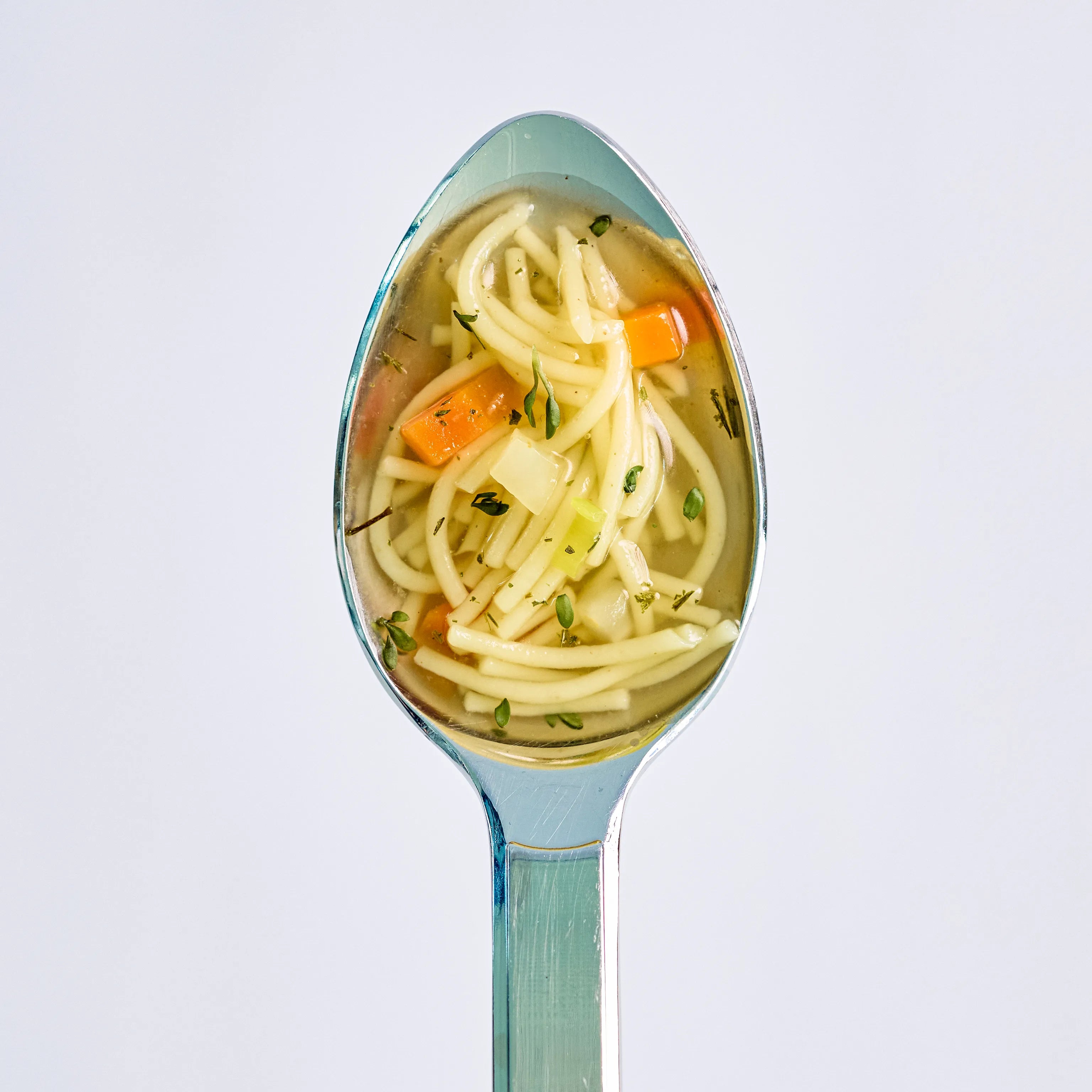 Löffel mit klarer Nudelsuppe, Gemüsestücken und Kräutern vor hellem Hintergrund, Nahaufnahme eines servierten Snacks