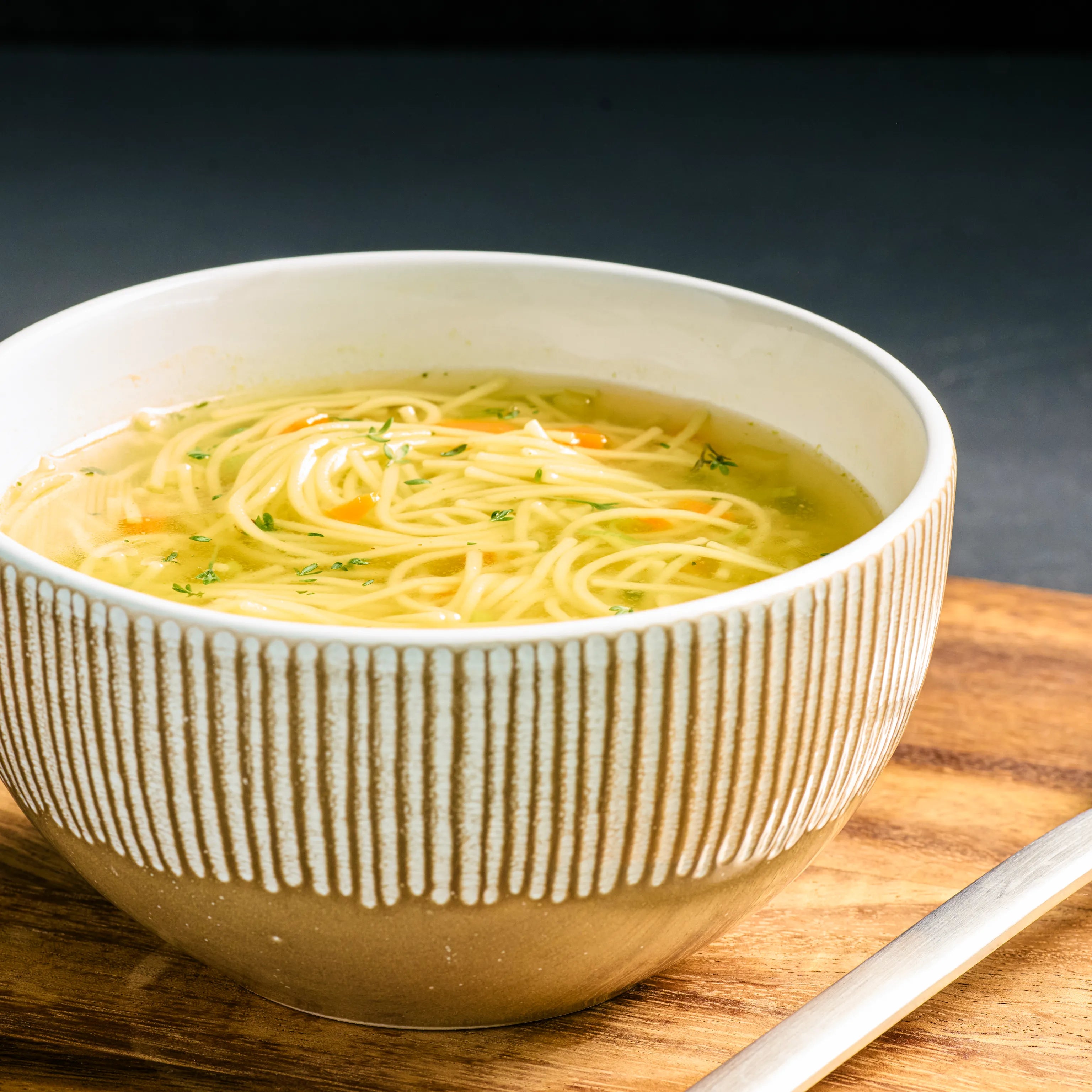Schüssel mit klarer Nudelsuppe auf Holzbrett, mit feinen Kräutern und Gemüsestücken, servierfertig angerichtet