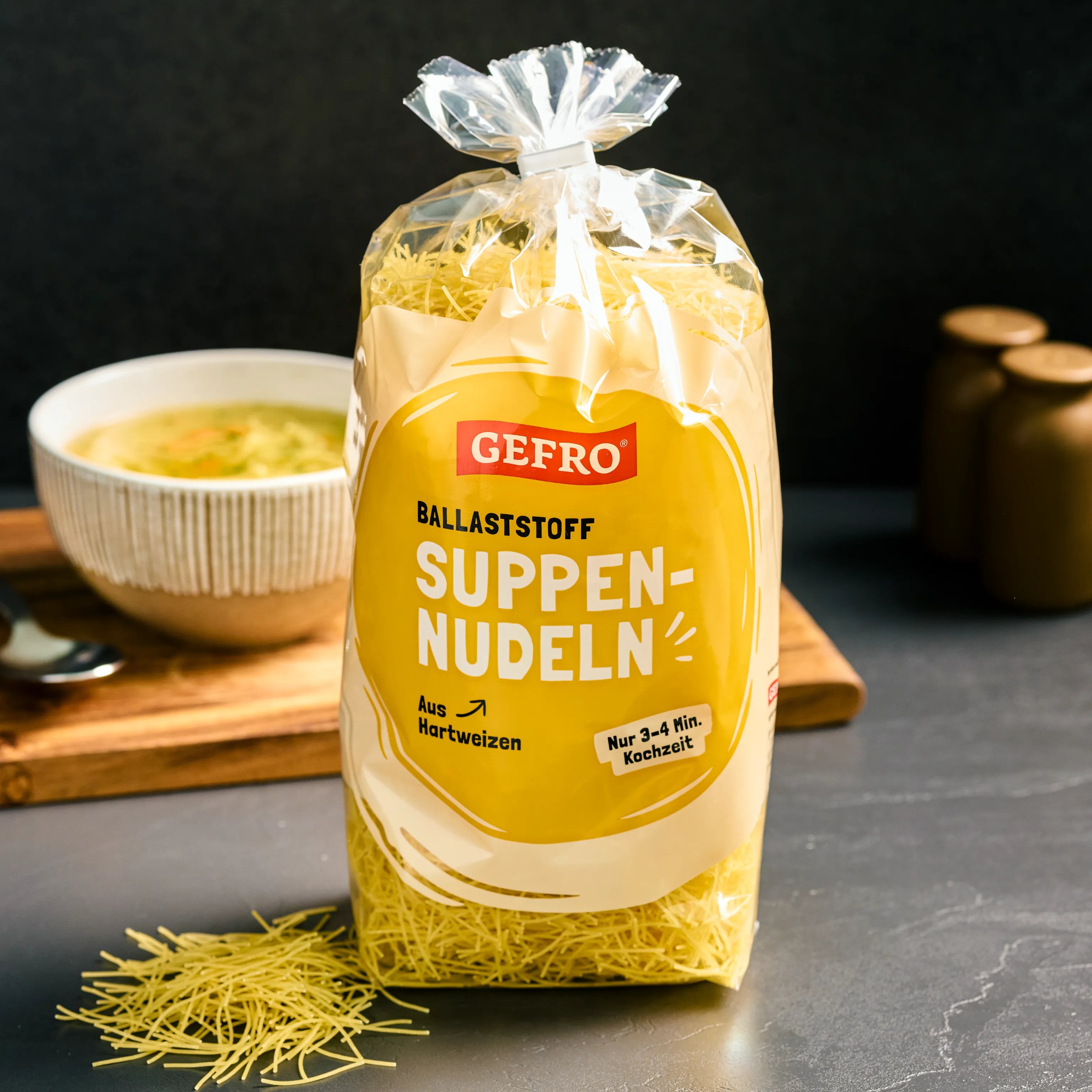 Packung GEFRO Ballaststoff Suppennudeln aus Hartweizen, daneben eine Schale Suppe auf Holzbrett