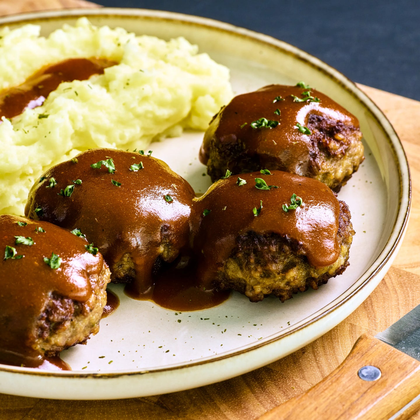 Fünf Frikadellen mit brauner Soße und Kartoffelpüree auf einem Teller, mit etwas Petersilie garniert
