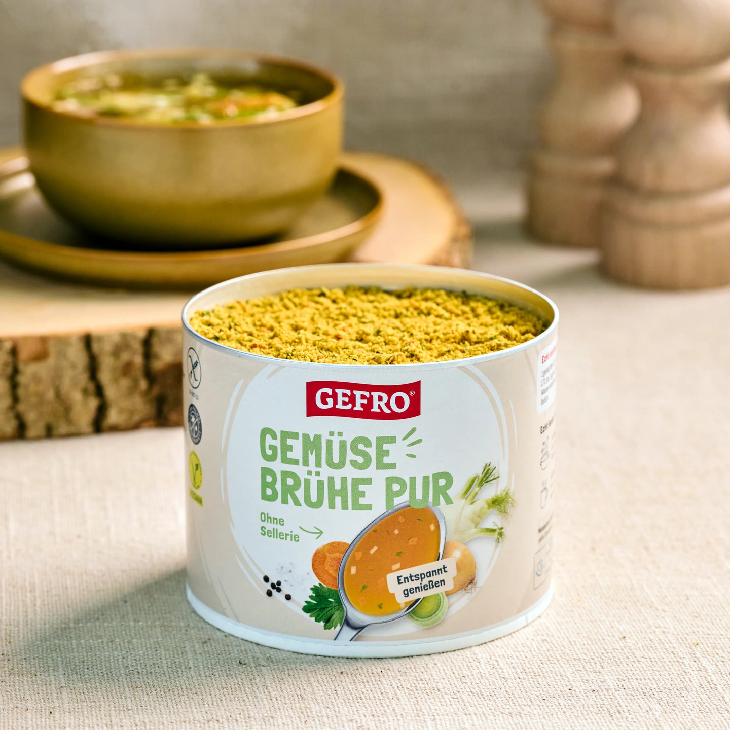 Dose GEFRO Gemüsebrühe Pur mit geöffnetem Pulverinhalt auf Tisch, dahinter eine Schüssel mit fertiger Suppe.