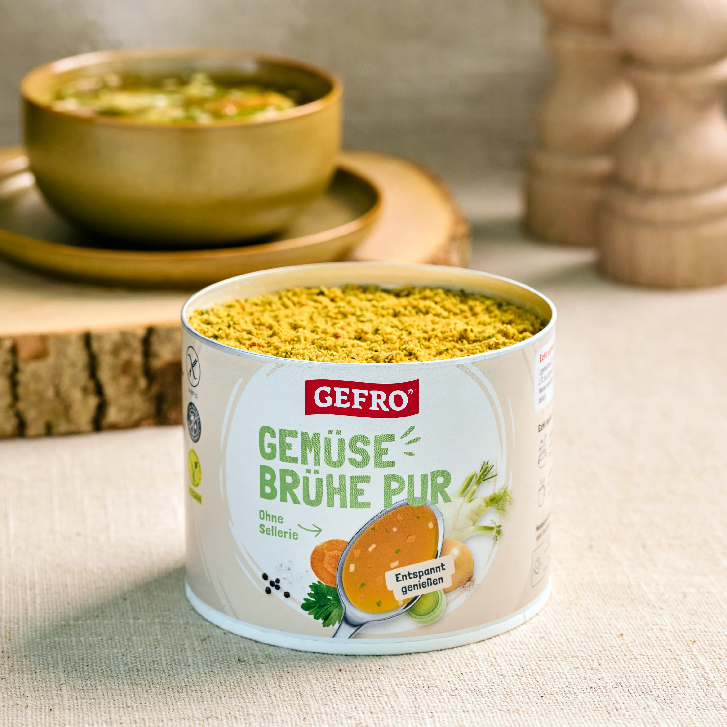 Dose GEFRO Gemüsebrühe Pur mit geöffnetem Pulverinhalt auf Tisch, dahinter eine Schüssel mit fertiger Suppe.