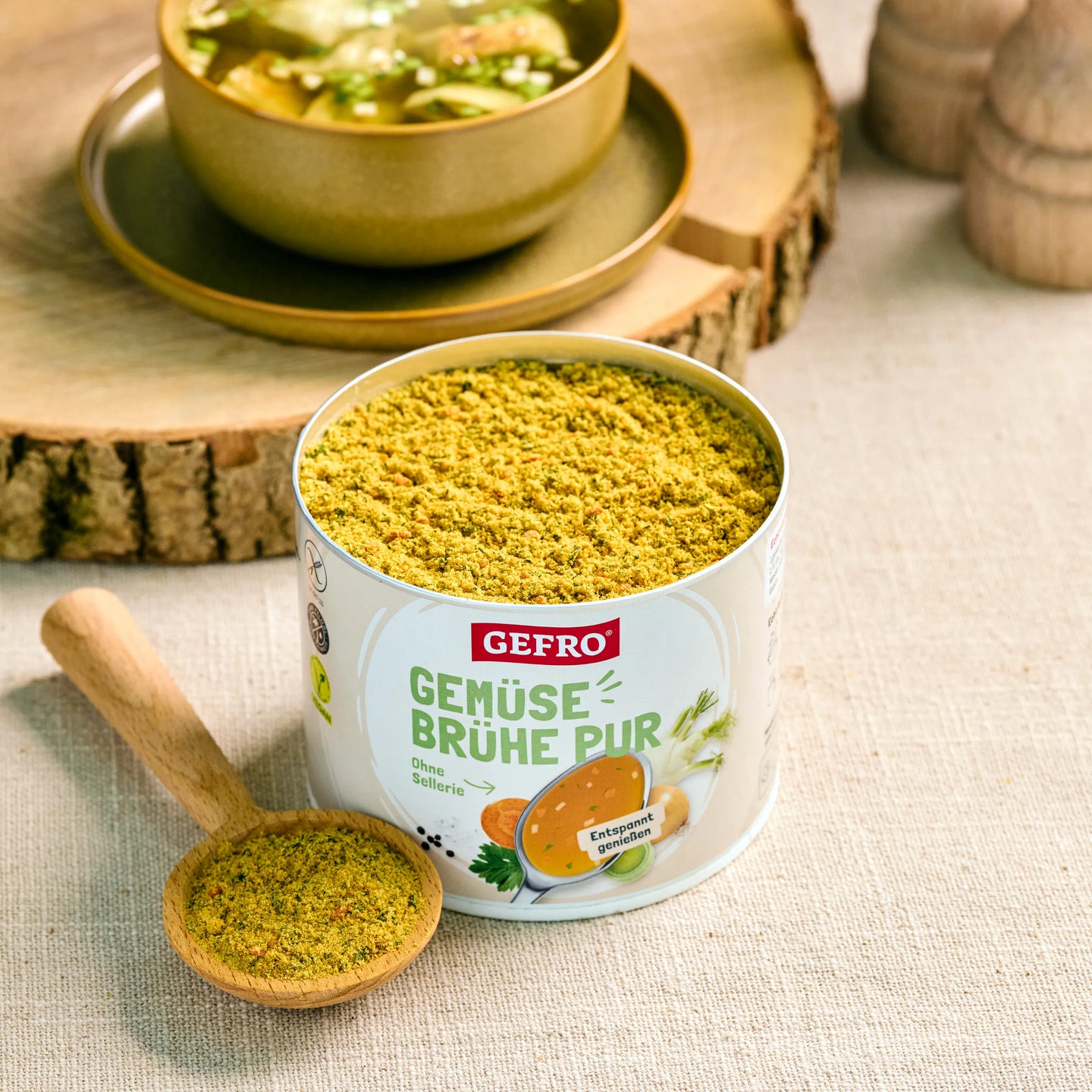 Dose GEFRO Gemüsebrühe Pur mit geöffnetem Pulver und Holzlöffel, dekoriert neben einer Schale Suppe.