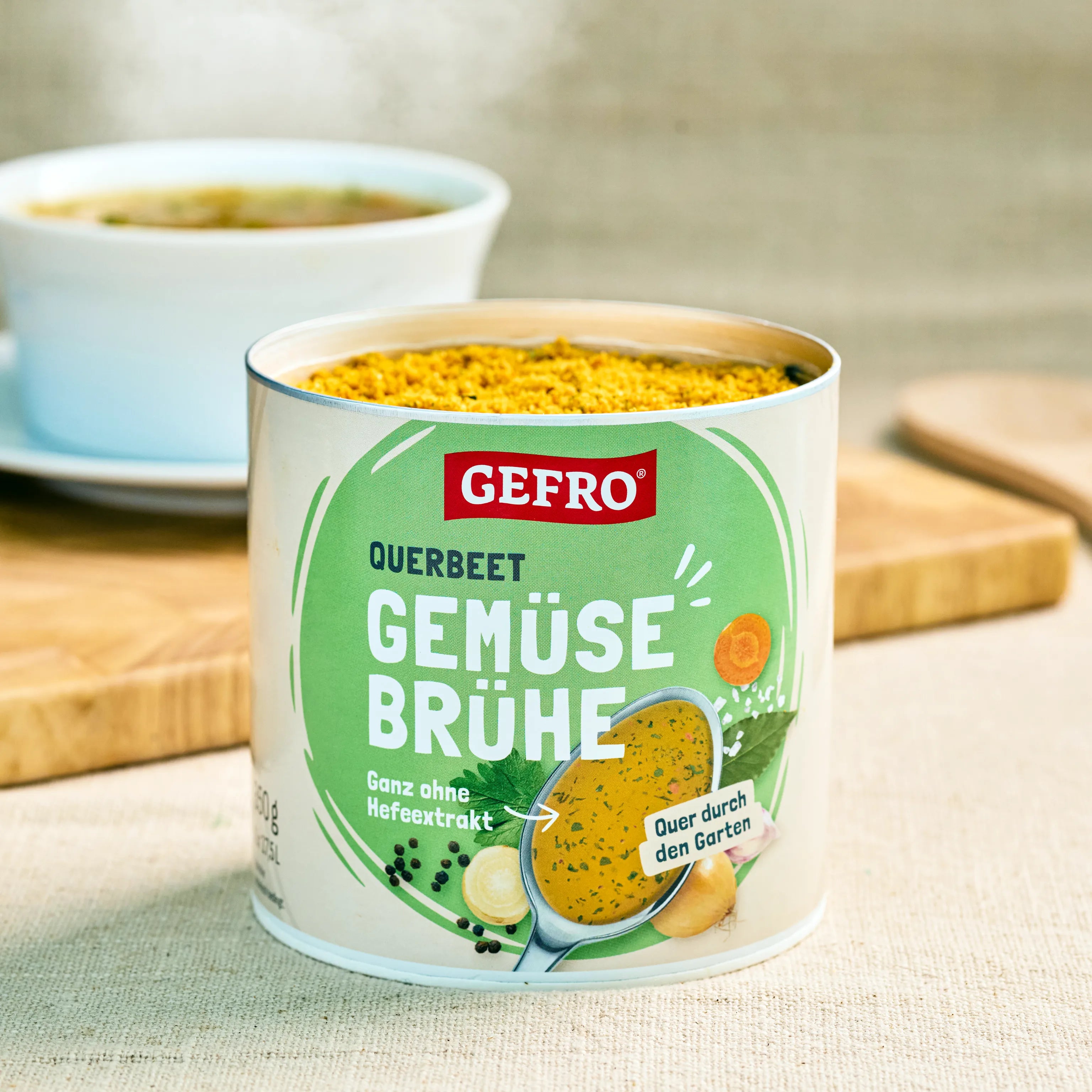 Offene Dose GEFRO Querbeet Gemüsebrühe auf Tisch, im Hintergrund eine Tasse mit zubereiteter Brühe.
