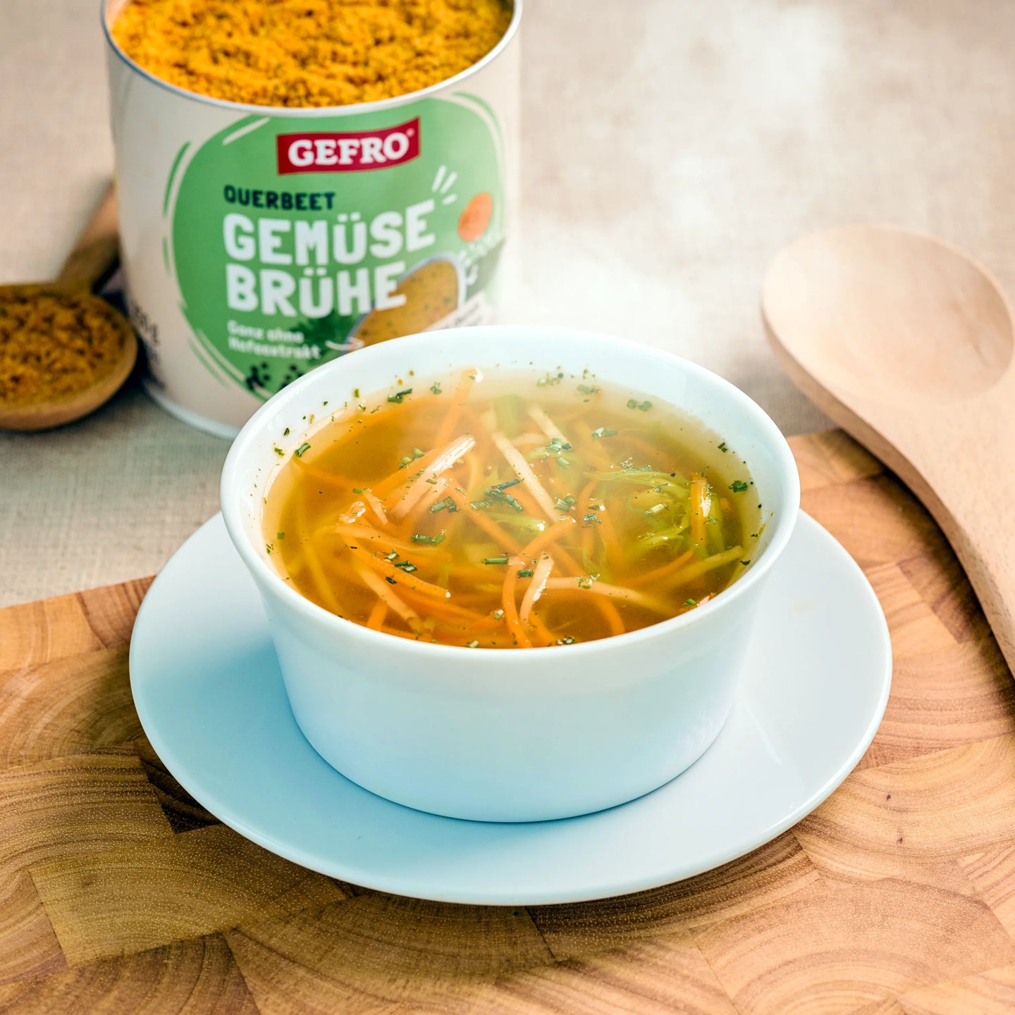Heiße Gemüsesuppe in weißer Schale, daneben Dose mit Gefro Querbeet Gemüsebrühe auf Holzbrett.