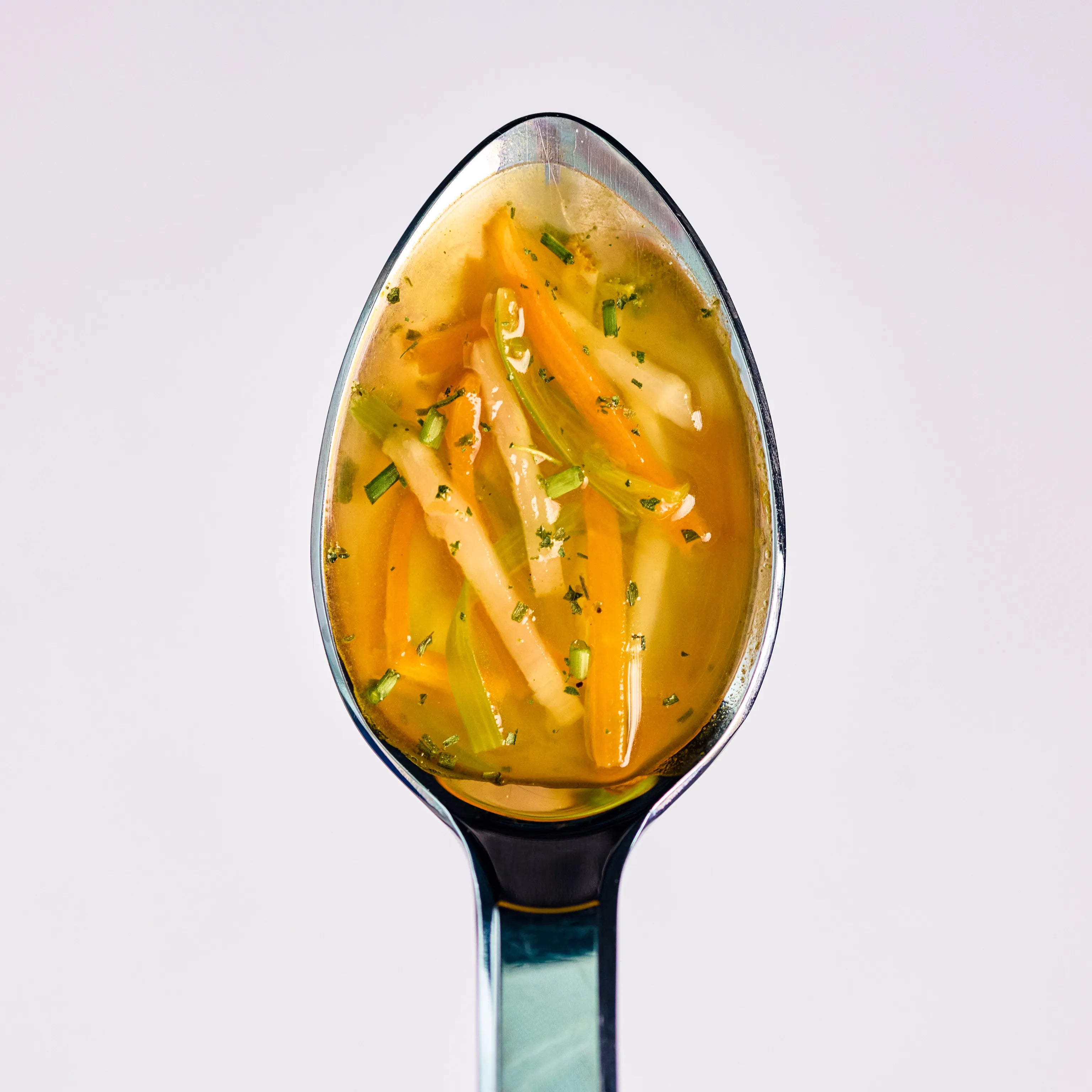 Löffel mit klarer Gemüsesuppe, feinen Karotten- und Selleriestreifen sowie Kräutern vor hellem Hintergrund