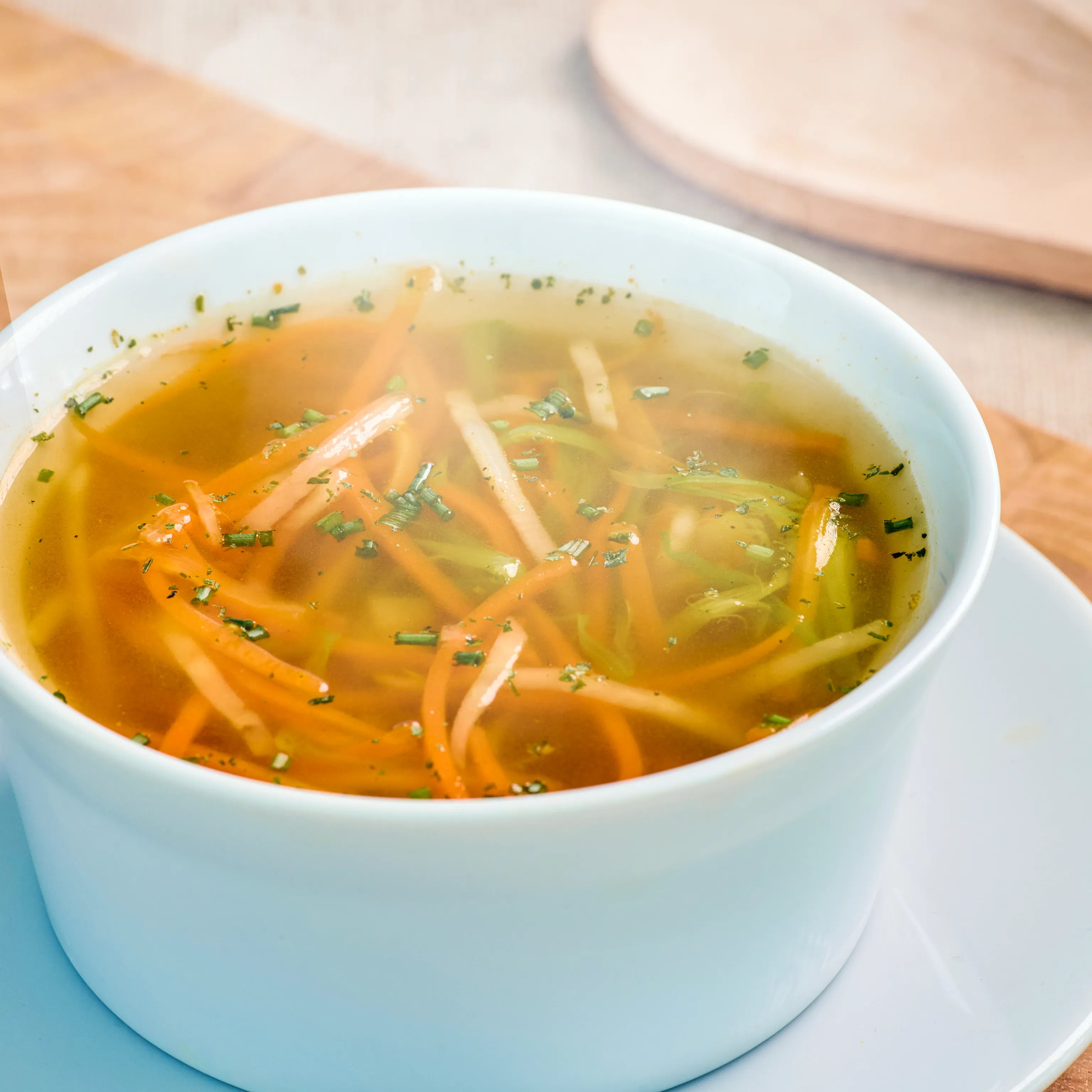 Weiße Schale mit klarer Gemüsesuppe, feinen Gemüsestreifen und Kräutern, serviert auf einem Unterteller.
