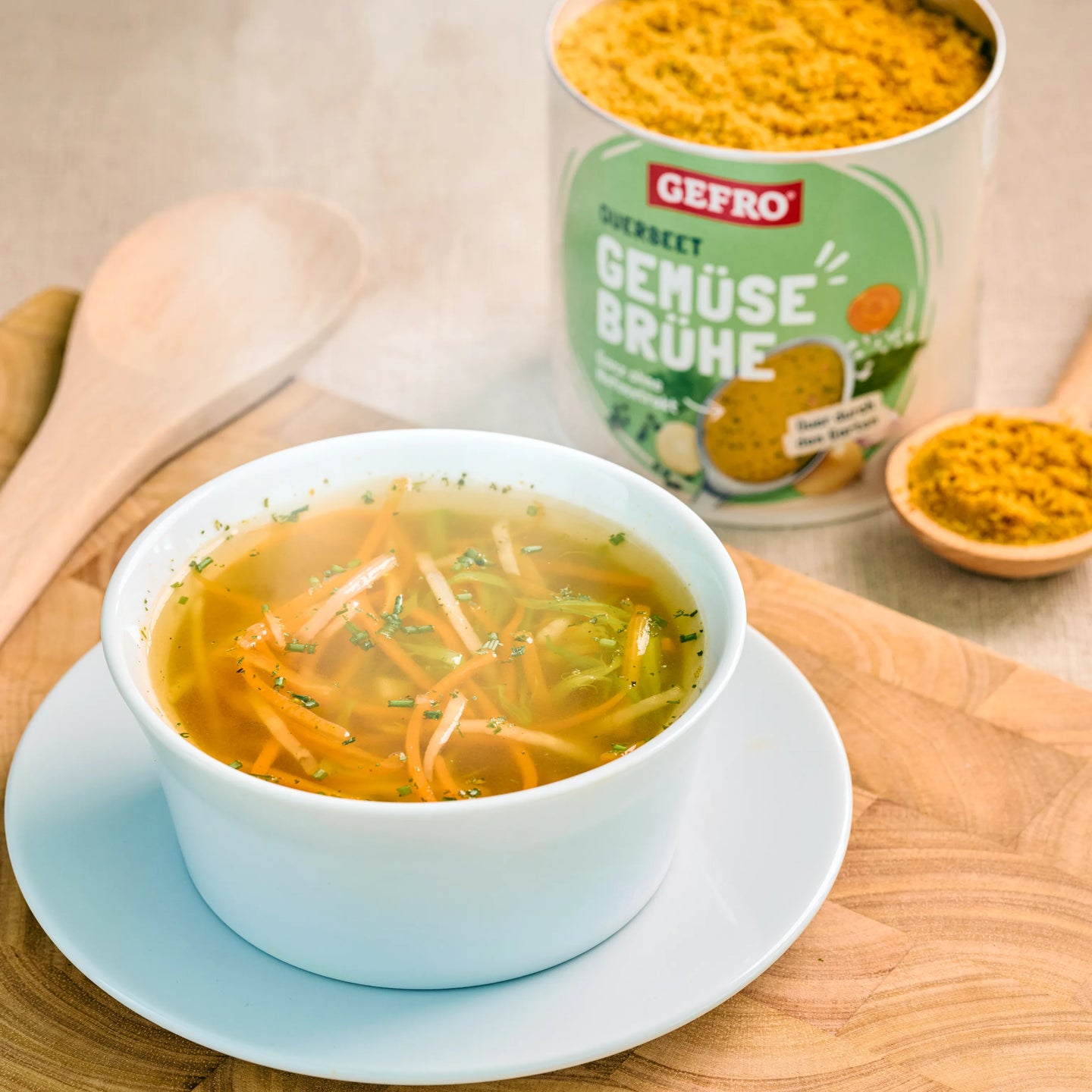 Schale mit heißer Gemüsesuppe auf Holzbrett, dahinter Dose GEFRO Gemüsebrühe und Löffel mit Brühepulver
