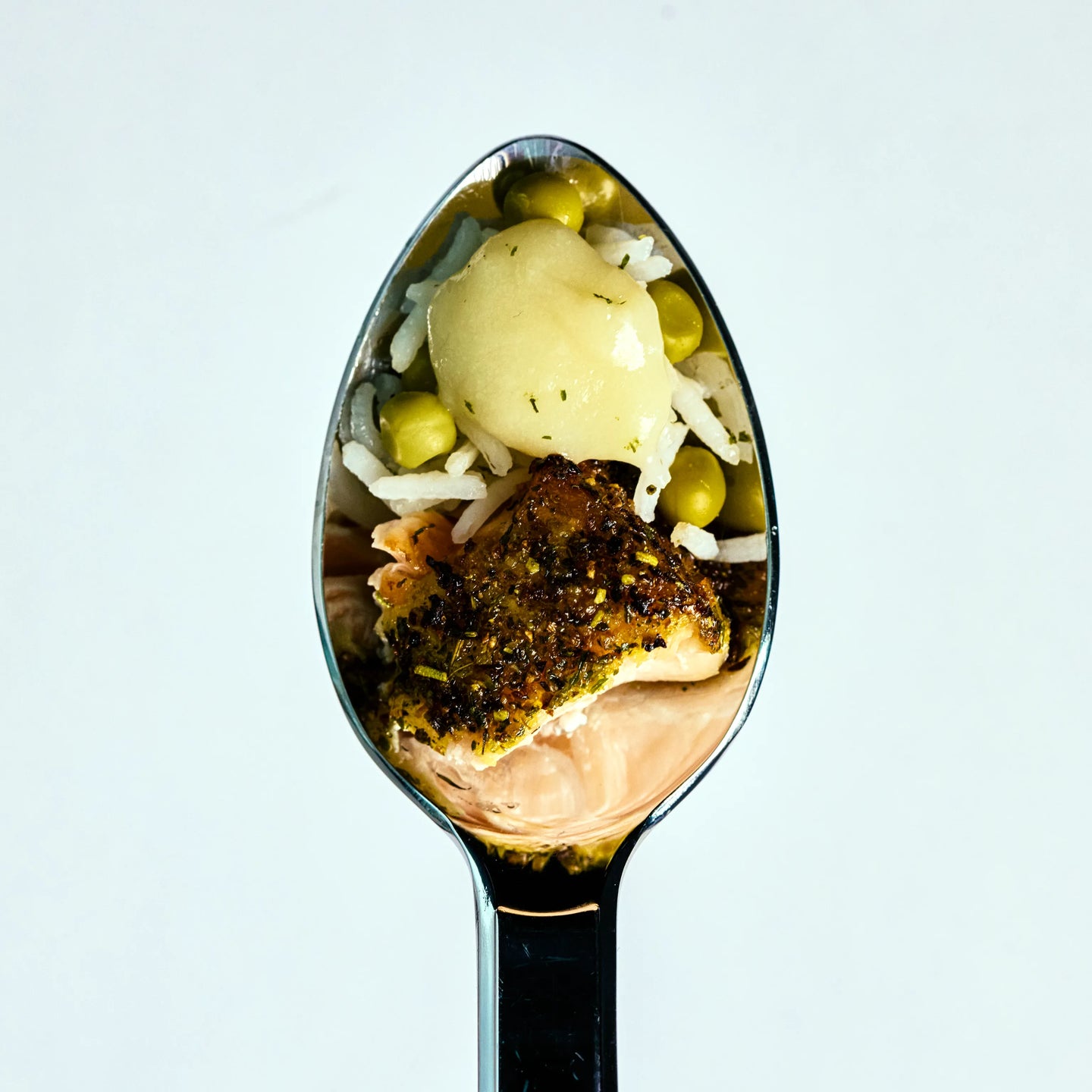Löffel mit Reis, Erbsen, einer kleinen Kartoffel und gebratenem Fischstück vor hellem Hintergrund