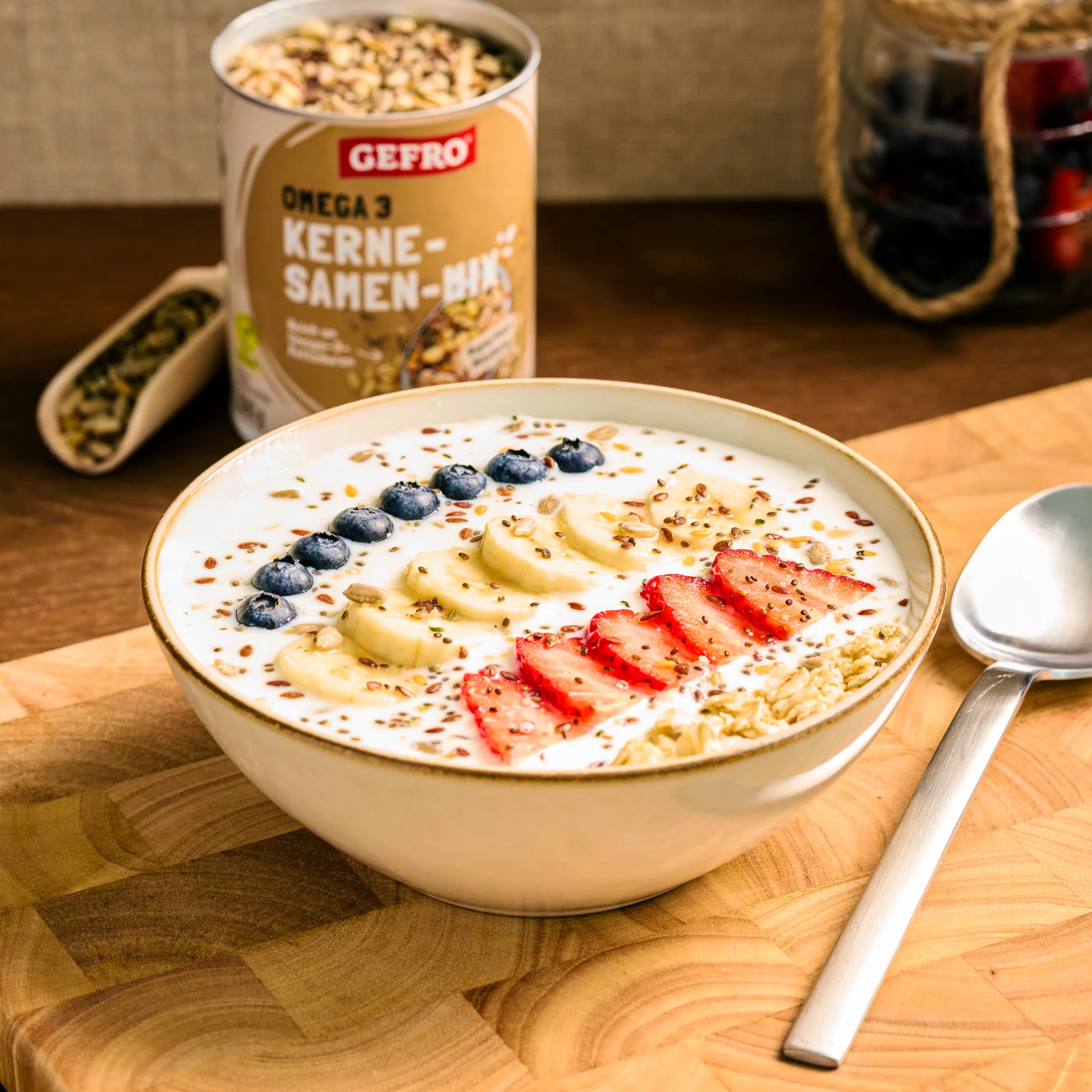 Schale mit Joghurt, Banane, Erdbeeren, Heidelbeeren und Körnern, daneben Löffel und Dose mit Kern-Samen-Mix