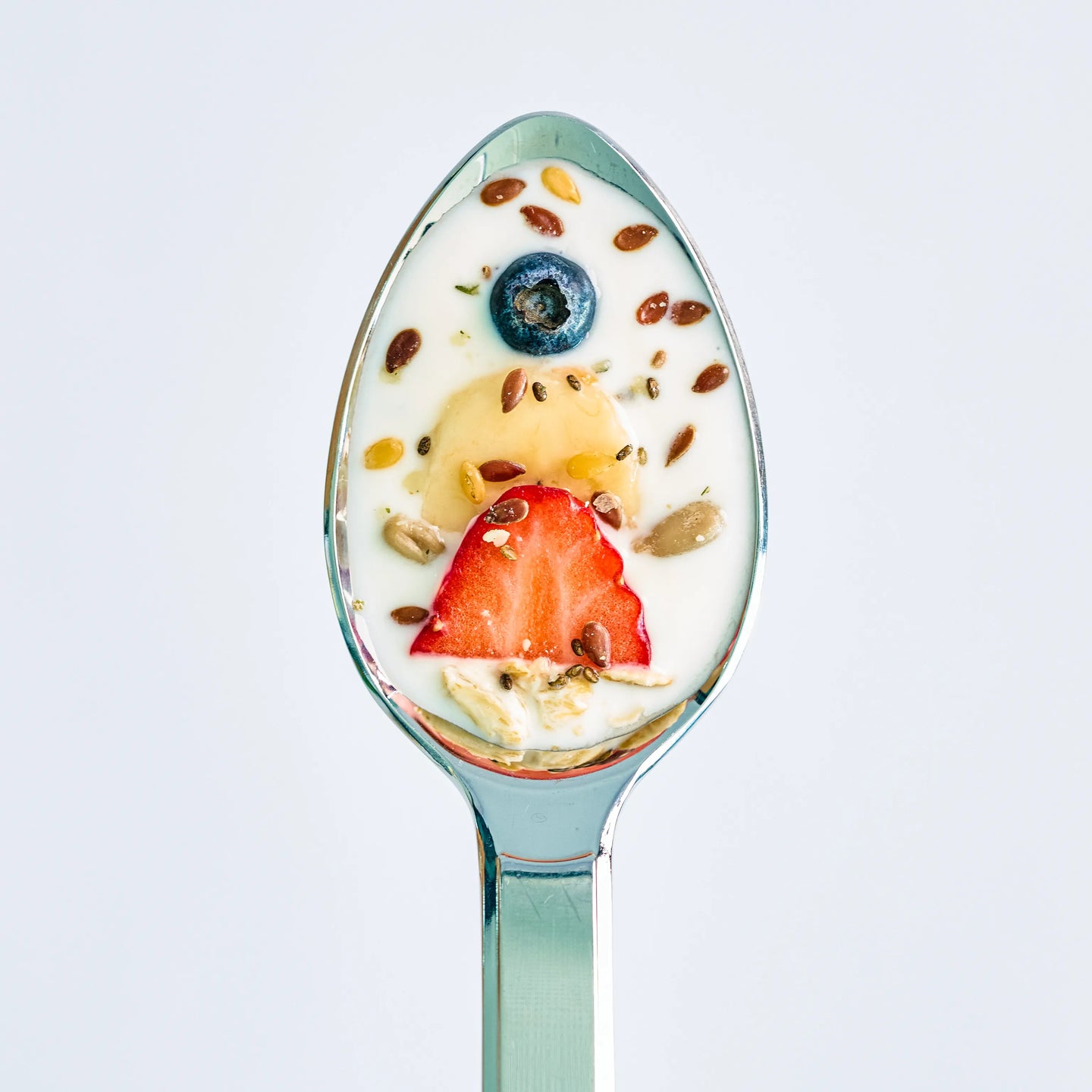 Löffel mit Joghurt, Erdbeerscheibe, Blaubeere, Bananenstücken und Samen vor hellem Hintergrund, Nahaufnahme