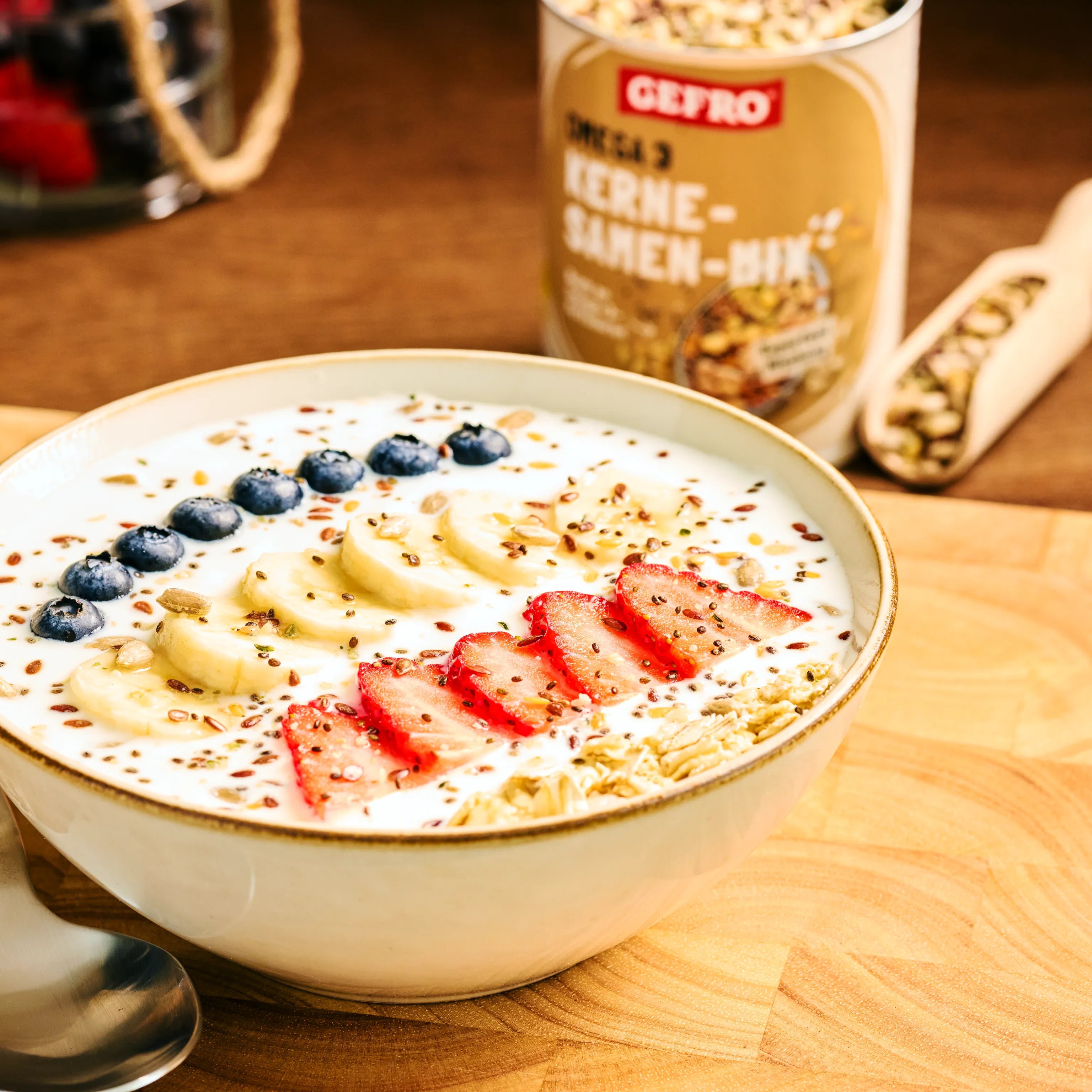 Schale mit Joghurt, Haferflocken, Banane, Erdbeeren, Blaubeeren und Saaten, dahinter Kerne-Samen-Mix von Gefro