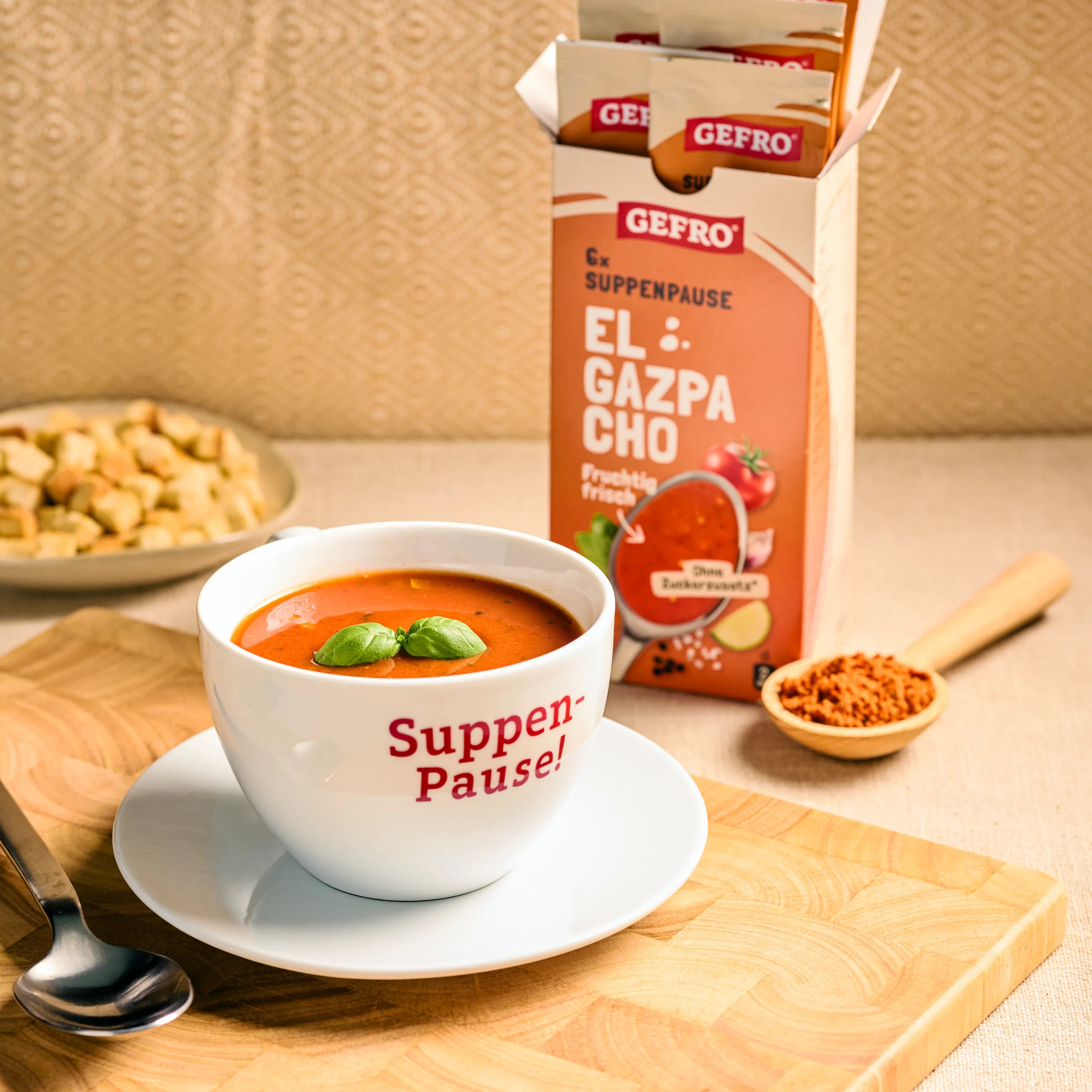 Tasse mit roter Gazpacho auf Unterteller vor geöffneter GEFRO-Packung, daneben Löffel und Gewürze auf Holzbrett